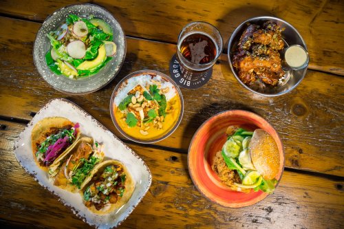 A vibrant spread of tacos, salad, wings, and a burger, accompanied by a glass of beer on a rustic wooden table.