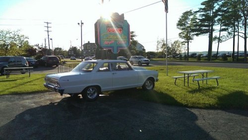 A classic white car parked on grass near a diner sign, with trees and a clear sky in the background.