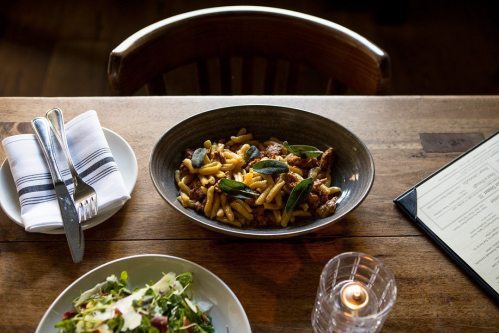 A bowl of pasta with herbs sits on a wooden table next to a salad and a menu, with a candle in the background.