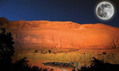 A boat glides on a river under a bright full moon, with a dramatic red rock formation illuminated at night.