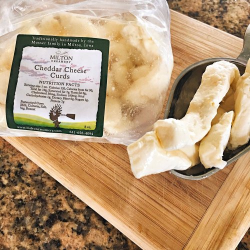 A bag of cheddar cheese curds on a wooden cutting board, with some curds in a measuring cup beside it.