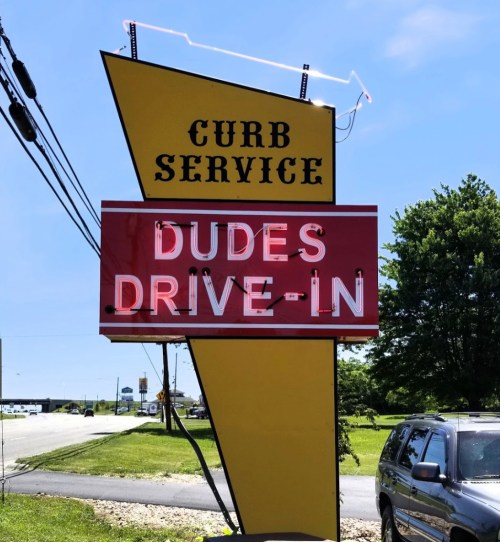 Brightly colored sign for "Dudes Drive-In" with "Curb Service" above, set against a clear blue sky.