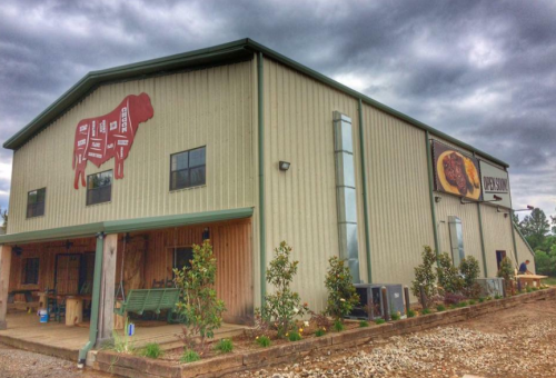 A large metal building with a cow silhouette sign, featuring wooden accents and a cloudy sky in the background.