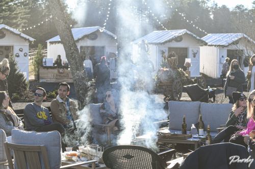 A cozy outdoor gathering with people sitting around a fire, smoke rising, and festive cabins in the background.