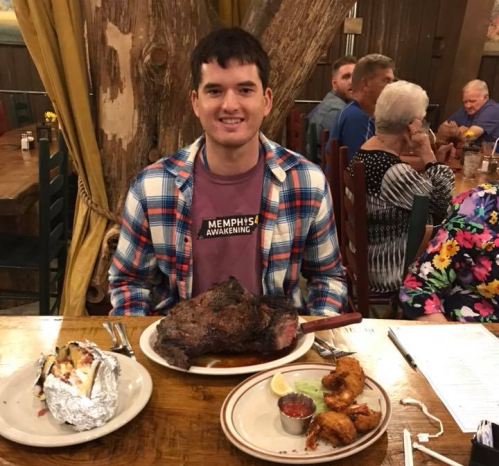 A man sits at a table with a large plate of meat, a baked potato, and shrimp, surrounded by diners in a rustic restaurant.