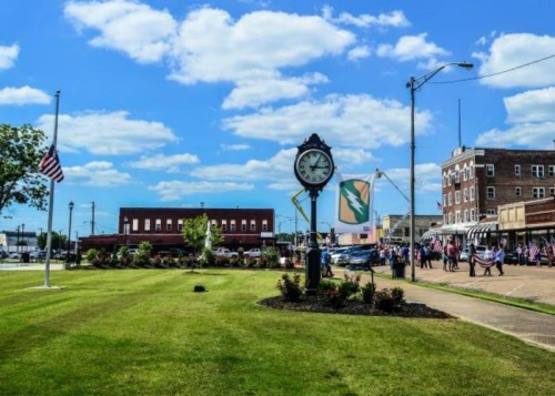 A town square with a clock, flags, and people gathered, set against a bright blue sky with fluffy clouds.