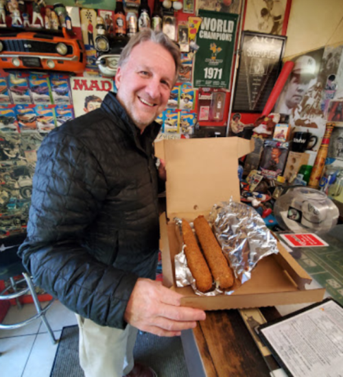 A smiling man holds a pizza box containing two large, wrapped food items in a colorful diner setting.