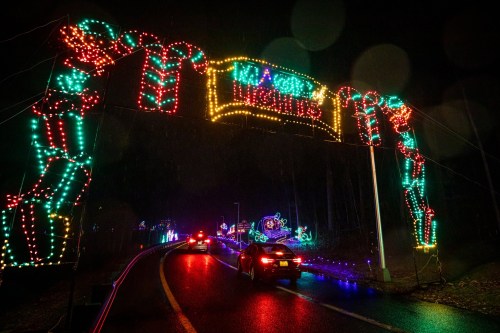 Colorful holiday lights arch over a road, with cars driving through a festive display on a rainy night.
