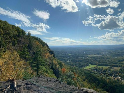 A scenic view from a mountain overlook, showcasing lush green valleys and a bright blue sky with scattered clouds.