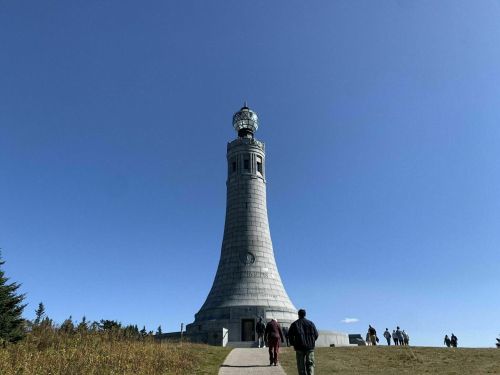 A tall stone lighthouse stands against a clear blue sky, with people walking towards it on a path.
