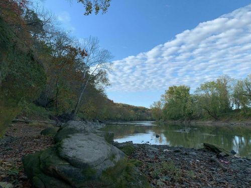 A serene river scene with rocky banks, trees, and a blue sky filled with clouds reflecting on the water's surface.