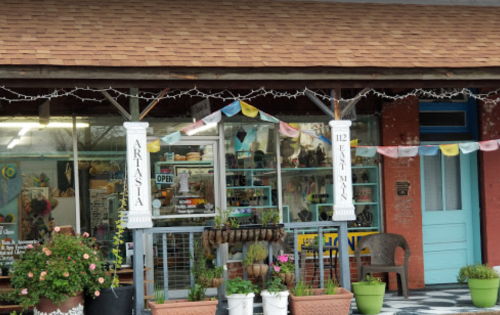 A quaint shopfront with colorful decorations, potted plants, and a sign reading "ARTASIA" and "112 East Main."