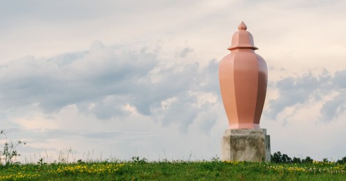 A large pink sculpture resembling an urn stands on a grassy hill under a cloudy sky.