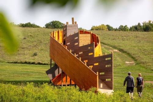 Two people walk towards a colorful, spiraling wooden structure on a grassy hillside.