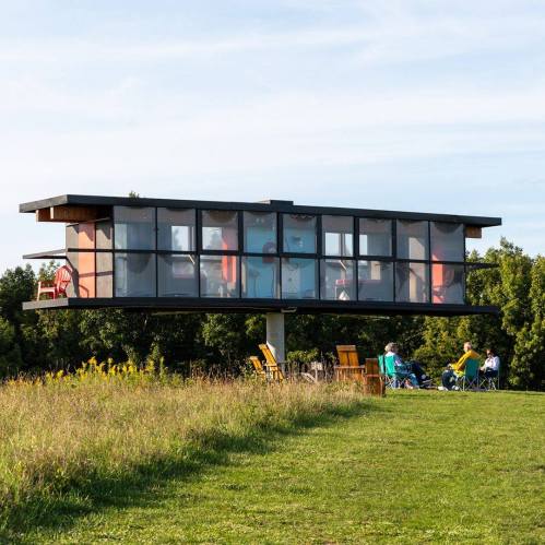 A modern glass house on stilts surrounded by greenery, with people relaxing on chairs nearby.