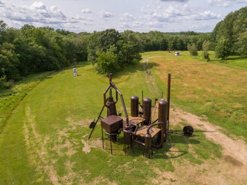 Aerial view of a large, industrial sculpture in a grassy field, surrounded by trees and a cloudy sky.