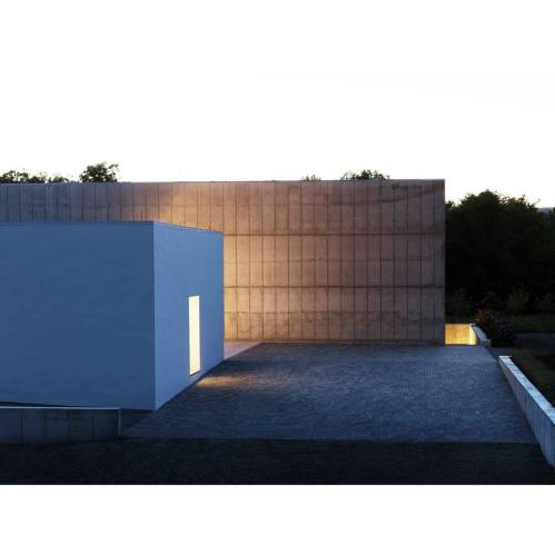 Modern architectural structure with a white cube and textured concrete wall, illuminated by soft light at dusk.