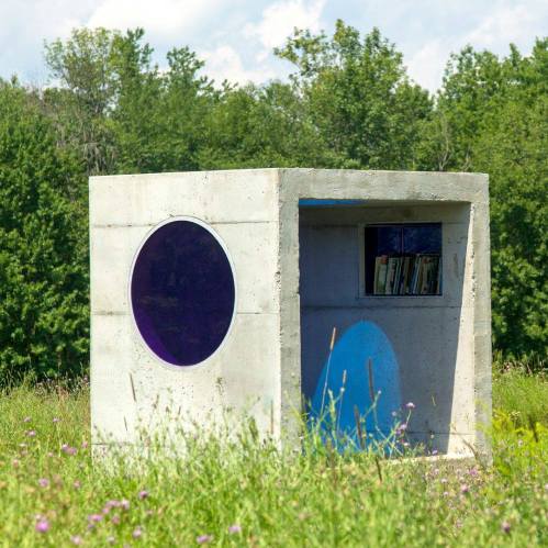 A modern concrete structure with a circular window, surrounded by wildflowers and trees in a sunny field.