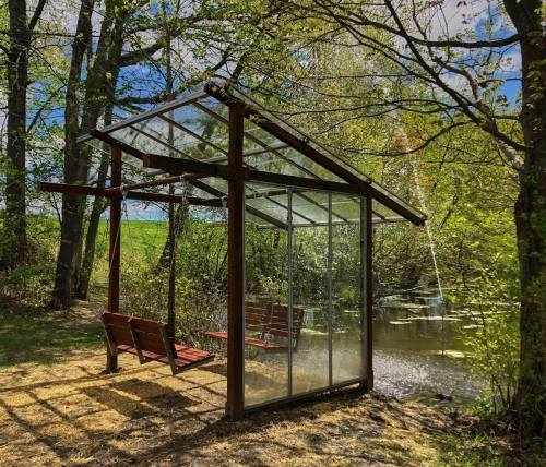 A glass-walled shelter with wooden swings beside a serene pond, surrounded by trees and greenery.