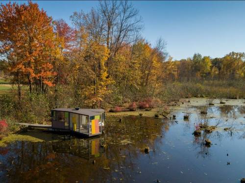 A modern cabin on a serene pond, surrounded by vibrant autumn foliage and clear blue skies.