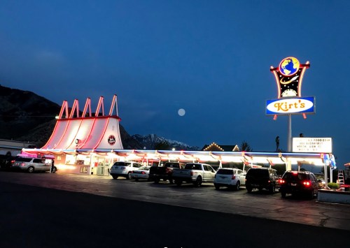 A brightly lit diner named Kirt's at dusk, with a mountain backdrop and a full moon in the sky.