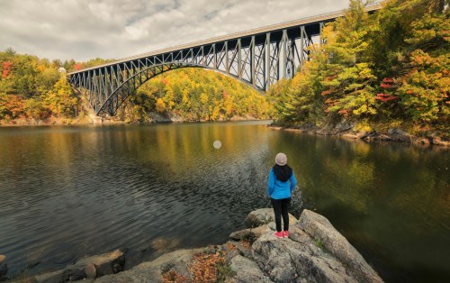 A person in a blue jacket stands on a rock by a lake, gazing at a large bridge surrounded by autumn foliage.