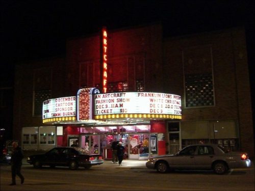 A vintage theater marquee at night, displaying event details and illuminated signage. Cars parked outside.