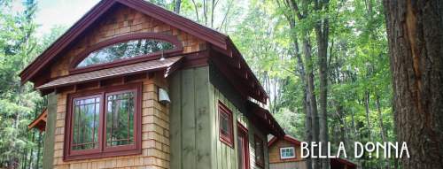 A charming wooden cabin nestled among trees, featuring large windows and a welcoming design. Text reads "Bella Donna."
