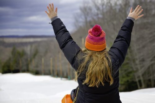 A person with long hair wearing a colorful hat raises their arms in celebration on a snowy slope.