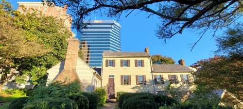 Historic house surrounded by greenery, with a modern skyscraper in the background under a clear blue sky.