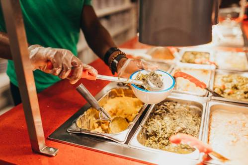 A person in gloves serves food from a buffet, using a ladle to scoop greens into a small bowl.