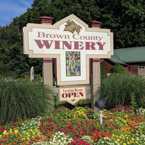 Sign for Brown County Winery with "Tasting Room OPEN" surrounded by colorful flowers and greenery.