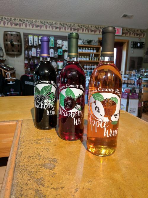 Three bottles of fruit wines: blackberry, strawberry, and apple, displayed on a wooden counter in a shop.