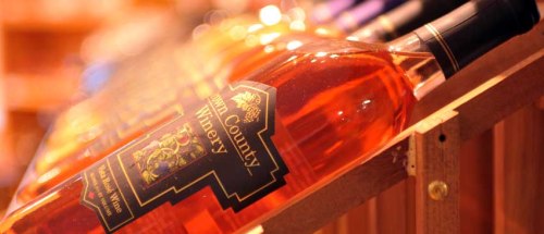 A close-up of a bottle of rosé wine on a wooden rack, with other bottles blurred in the background.