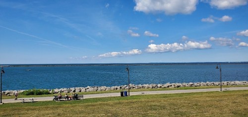 A serene waterfront view with a clear blue sky, rocky shoreline, and people sitting on benches.