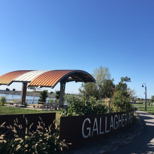 A park entrance sign reading "Gallagher Park" with a pavilion and clear blue sky in the background.