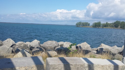A serene view of a blue lake with rocky shoreline and green trees under a partly cloudy sky.