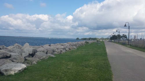 A scenic waterfront path lined with rocks and grass, under a partly cloudy sky.