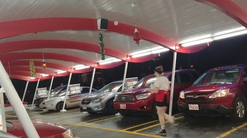 A drive-in restaurant with red awnings, several parked cars, and a staff member walking by.