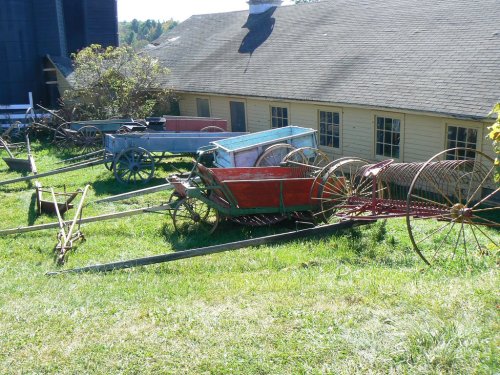 A collection of vintage farm wagons in various colors, resting on green grass near a yellow building.