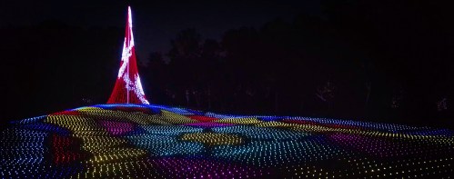 A colorful light display on a hillside at night, featuring a tall illuminated structure resembling a tree.