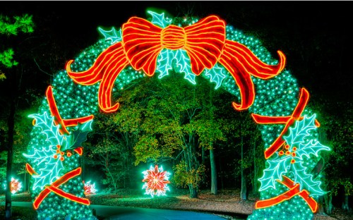 A brightly lit archway decorated with a red bow and holly, surrounded by colorful holiday lights in a wooded area.