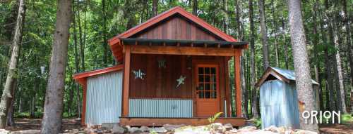 A small wooden cabin with a porch, surrounded by trees, and a shed nearby.