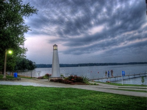 A lighthouse stands by a lake at dusk, with people walking along the shore under a cloudy sky.
