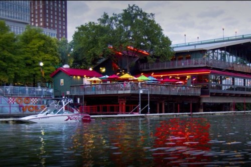 A waterfront restaurant with colorful umbrellas, surrounded by trees and city buildings, with a boat docked nearby.
