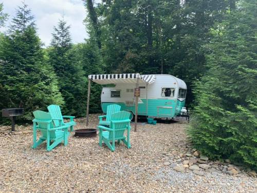 A vintage camper surrounded by trees, with turquoise chairs and a fire pit on a gravel area.