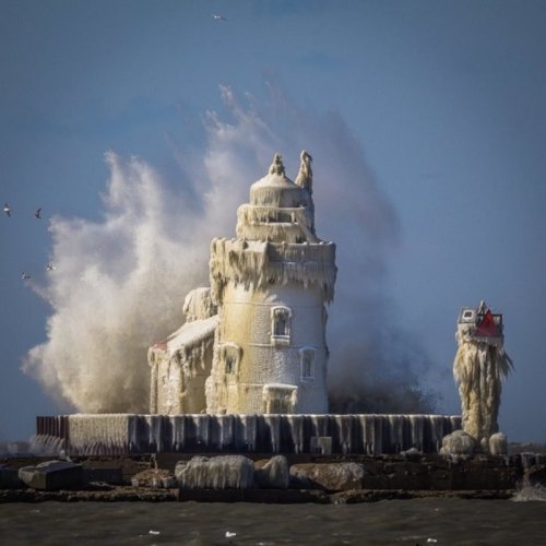 A lighthouse covered in ice and snow, with waves crashing against it, creating a dramatic winter scene.