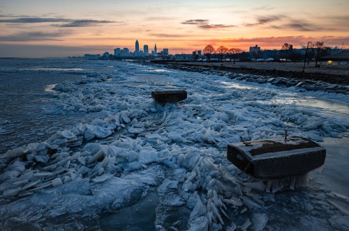 Sunset over a frozen lake with ice formations and a city skyline in the background.