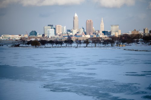 A snowy landscape with a frozen lake in the foreground and a city skyline in the background under a cloudy sky.