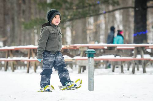 A young boy in winter clothing walks on snowshoes in a snowy park with trees and picnic tables in the background.